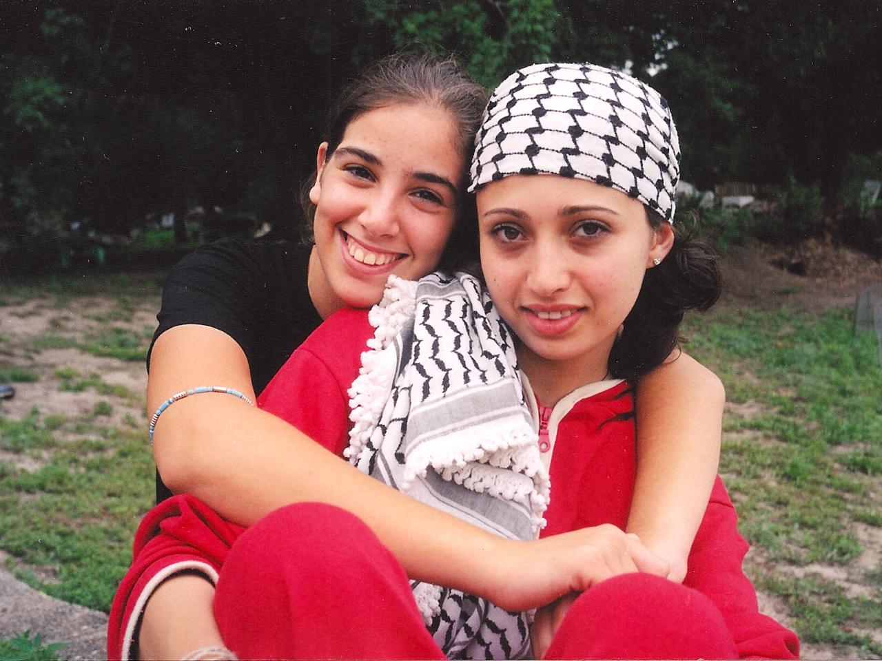 A Palesintian teen girl with a keffiyeh around her head sits on the grass. Behind her, and with arms wrapped around her, is an Israeli teen girl. They both smile for the camera.